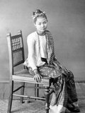 The young woman wears a striped <i>hta-mein</i> (wrap-around skirt) and a close-fitting jacket of fine muslin or cotton known as an <i>ein-gyi</i>. In her hair she wears a floral headband and she is adorned with necklaces, earrings, bracelets and rings.<br/><br/>

During the Konbaung Dynasty (1752-1885), rich jewellery, fine fabrics such as silk and garments such as her jacket were reserved for court officials and their wives by law. After the fall of the Burmese monarchy in 1885 they were worn by the wealthy.<br/><br/>

This photograph is from an album recording aspects of Lord Elgin's Burma tour of November to December 1898. Victor Alexander Bruce (1849-1917), 9th Earl of Elgin and 13th Earl of Kincardine, served as Viceroy of India between 1894 and 1899.